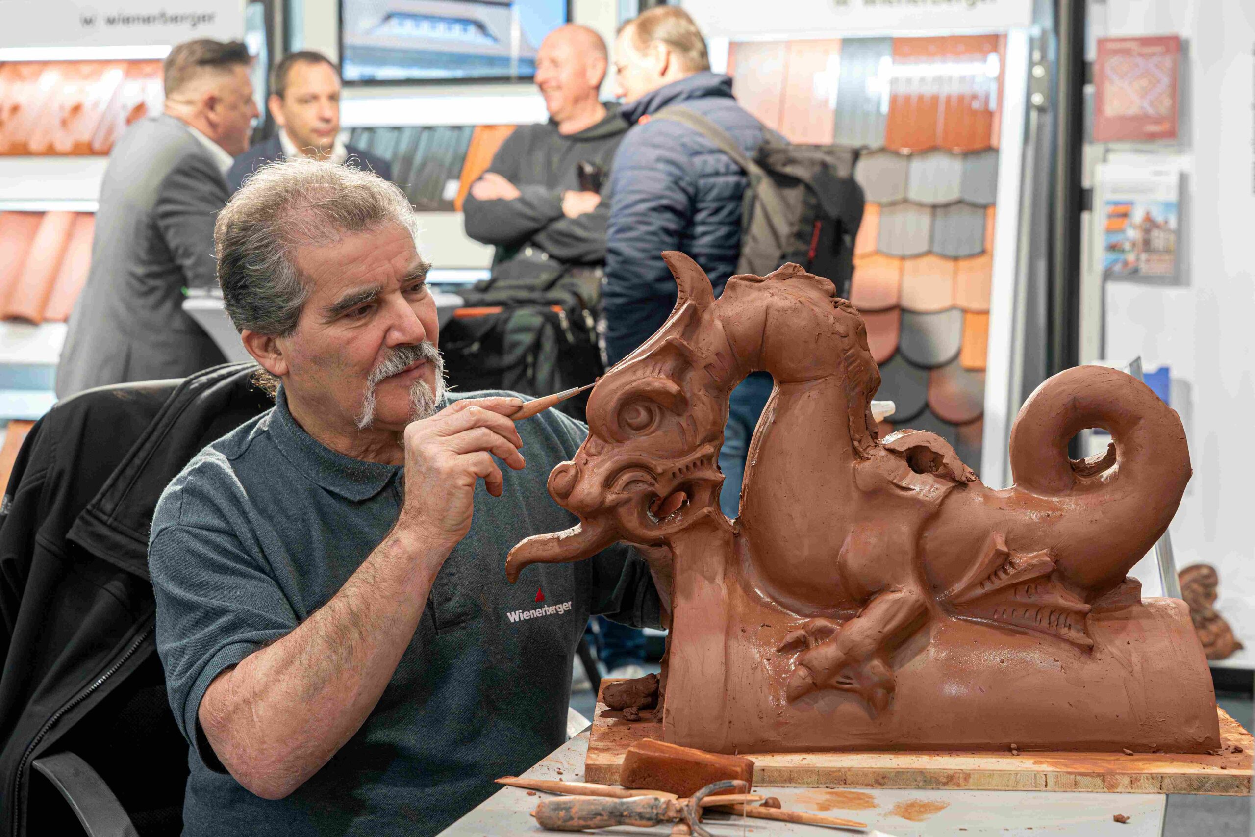 Eine Person modelliert mit einem Werkzeug eine detailreiche Tonfigur in Drachenform auf einem Tisch; im Hintergrund stehen mehrere Personen an einem Messestand mit Dachziegelmustern.