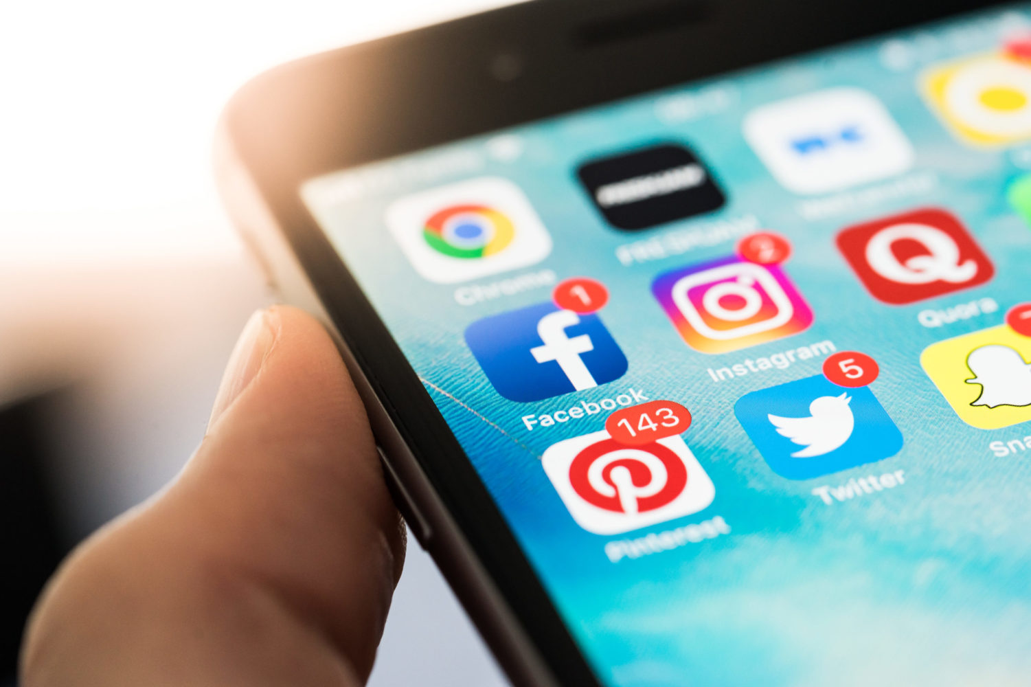 Smartphone in einer Hand mit geöffnetem Startbildschirm. Sichtbar sind mehrere Social-Media-Apps, darunter Facebook, Instagram, Pinterest, Twitter und Snapchat, teilweise mit Benachrichtigungsanzeigen.